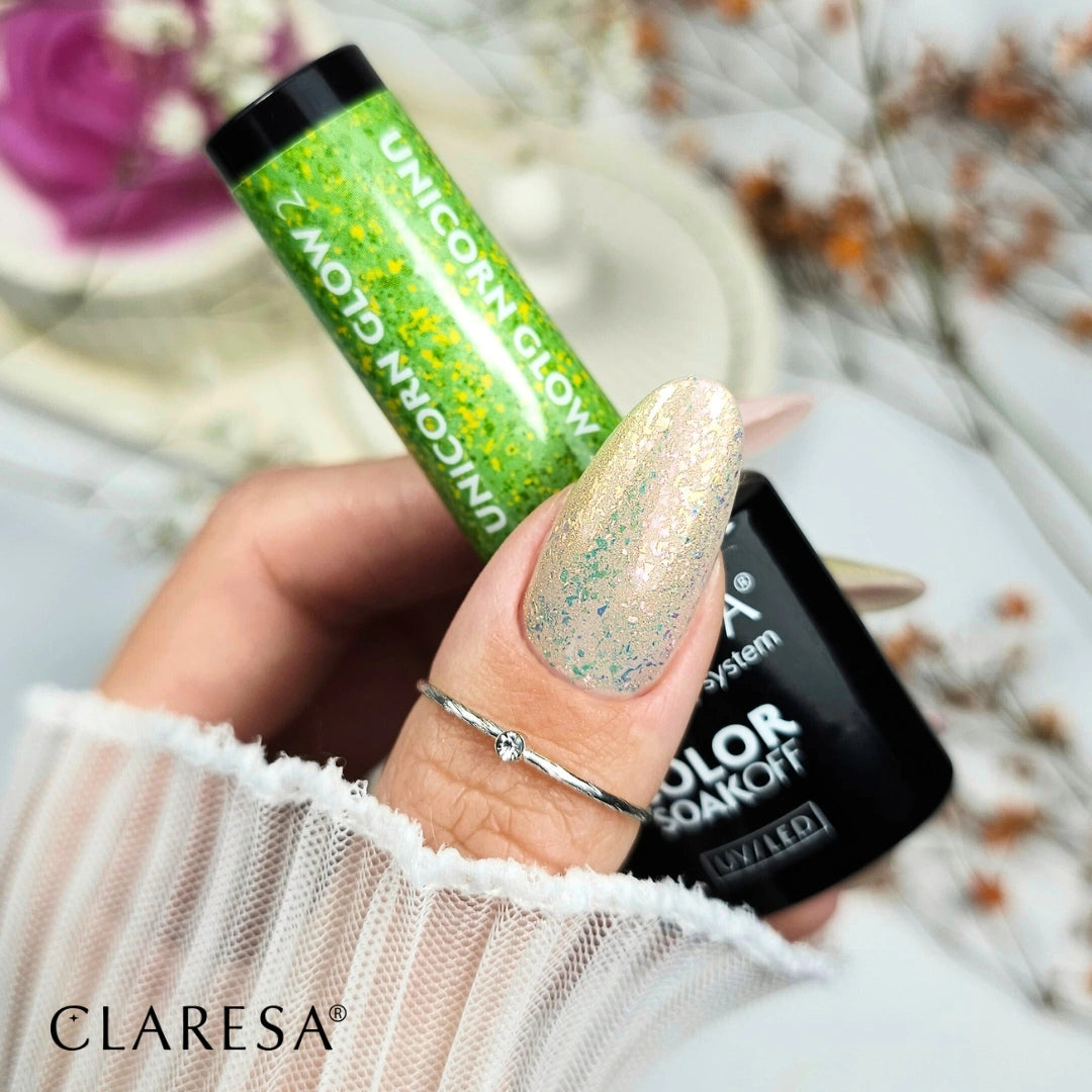 Claresa prekės ženklo 'Unicorn Glow' gelinis nagų lakas žalioje pakuotėje su blizgučiais, tepamas ant nago su subtiliu spalvų perėjimu. Ranka su žiedu laiko laką, fone – baltas audinys ir dekoratyviniai elementa